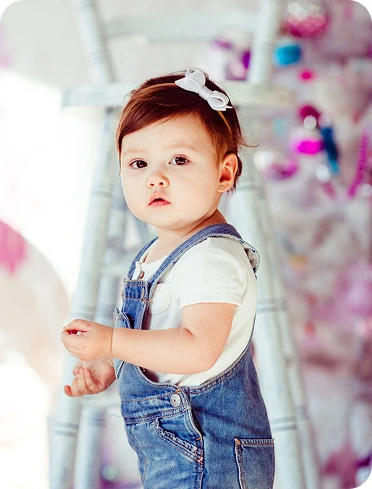 Young child wearing denim overalls and a white bow in a colourful playroom.