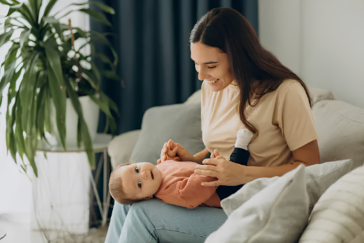 Mother sits on a light sofa, cradling a baby in a peach onesie and smiling warmly at the child—cozy home setting with green plant nearby.