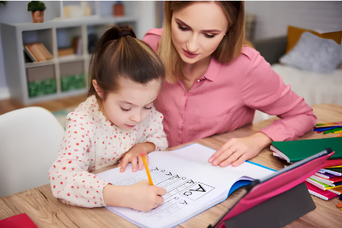 Mother-guiding daughter as she draws with a pencil at a wooden table, with notebooks and colorful markers nearby in a cozy room.