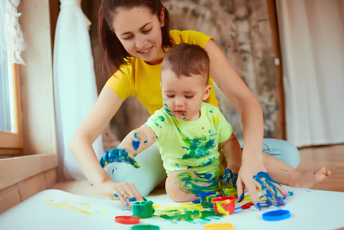 Mother helping a toddler paint with bright colors on a white table, both hands covered in blue, green, and yellow paint.